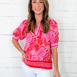 Woman wearing a pink and red patterned blouse with white pants against a white wooden background