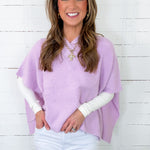Woman wearing a light purple poncho over a white shirt and pants, standing against a white wooden panel background.