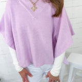 Person wearing a light purple poncho over a white shirt and pants, standing against a white wooden panel background.