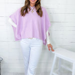 Woman wearing a light purple poncho and white pants against a white wooden panel background