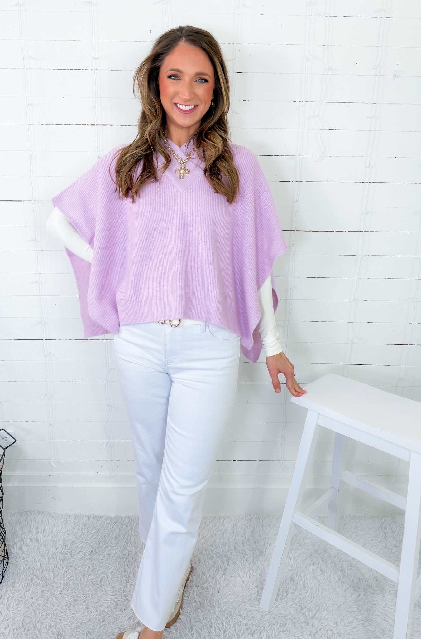 Woman wearing a light purple poncho and white pants against a white wooden panel background