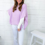 Woman wearing a light purple poncho over a white top and pants, standing against a white wooden paneled wall.