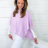 Woman wearing a light purple poncho over a white top and pants, sitting on a white stool against a white wooden panel background.