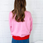 Person wearing a pink sweatshirt over a red shirt with blue jeans against a white wooden panel background