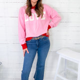 Woman wearing a pink sweatshirt with 'LOVE' on it, blue jeans, and gold shoes in a room with white wooden walls.