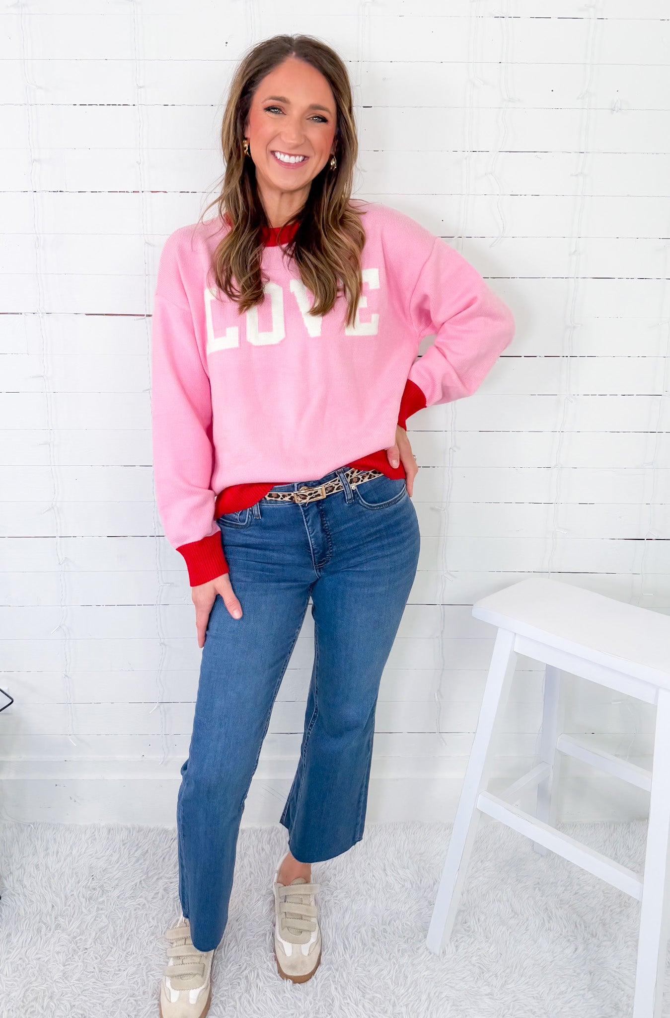 Woman wearing a pink sweatshirt with 'LOVE' on it, blue jeans, and gold shoes in a room with white wooden walls.