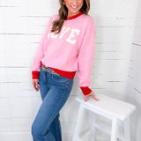 Woman wearing a pink sweatshirt with 'LOVE' on it, standing next to a white stool against a white wooden wall.