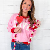 Woman wearing a pink sweatshirt with 'LOVE' holding a heart-shaped prop and a colorful necklace against a white wooden background.