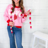 Woman wearing a pink sweatshirt with 'LOVE' and heart designs, holding a decorative garland against a white wooden wall.