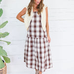 A woman modeling a brown and white buffalo plaid midi dress with a cream-colored quilted vest, standing in front of a white wood-paneled wall next to a potted green plant.