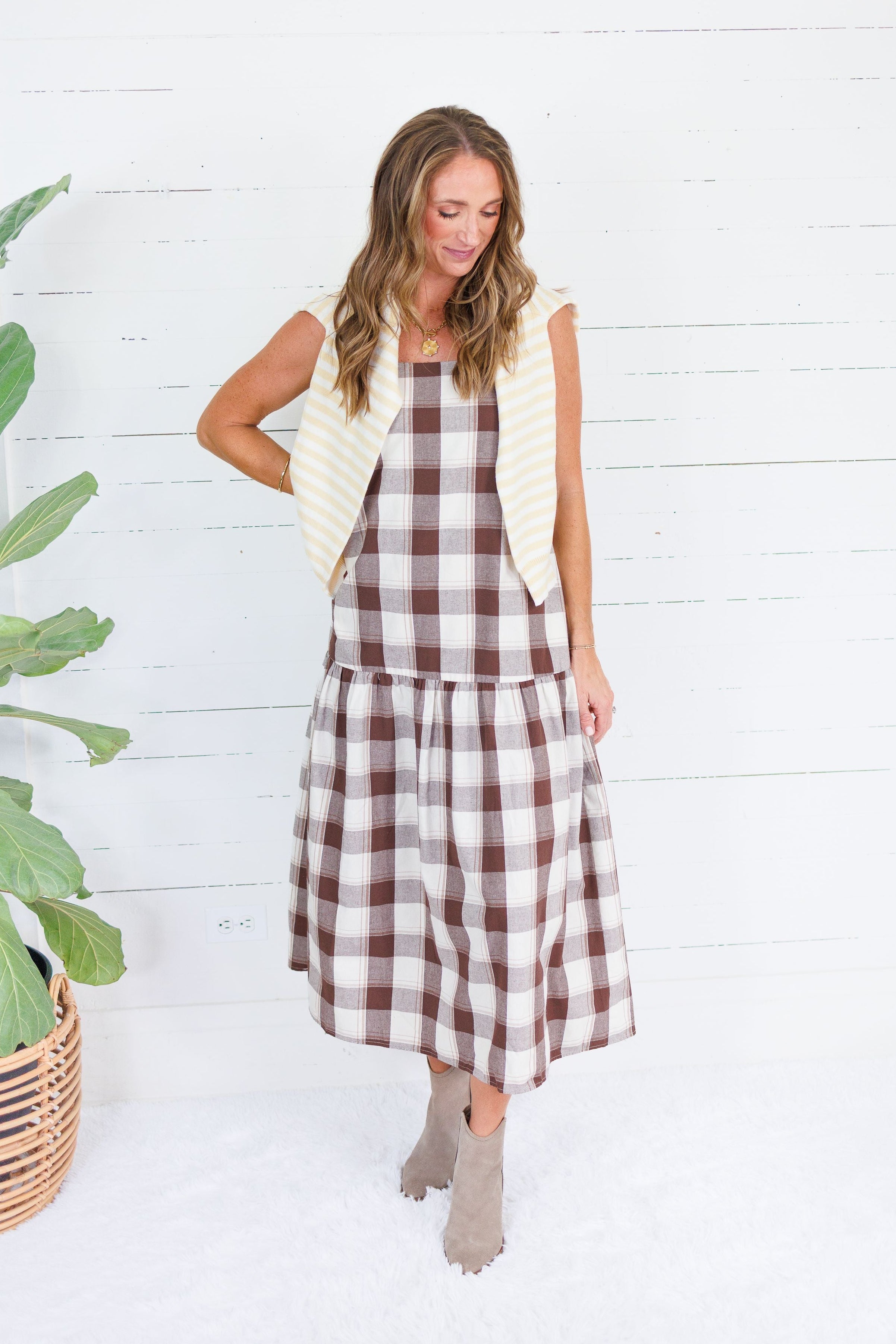 A woman modeling a brown and white buffalo plaid midi dress with a cream-colored quilted vest, standing in front of a white wood-paneled wall next to a potted green plant.