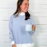 Woman wearing a blue striped sweater and white pants, holding a mug against a white wooden panel background.