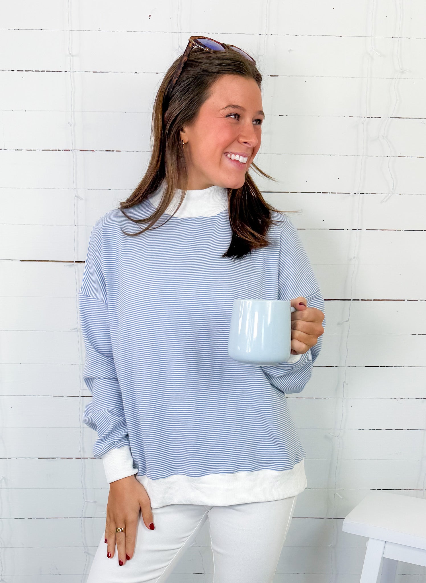 Woman wearing a blue striped sweater and white pants, holding a mug against a white wooden panel background.