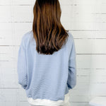 Person wearing a light blue sweater and white pants against a white wooden panel background