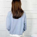 Person wearing a light blue sweater and white pants against a white wooden panel background