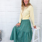 Woman wearing a yellow sweater and green patterned skirt sitting on a white stool against a white wooden paneled wall.
