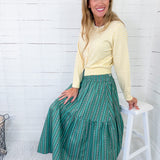 Woman wearing a yellow sweater and green patterned skirt sitting on a white stool against a white wooden paneled wall.