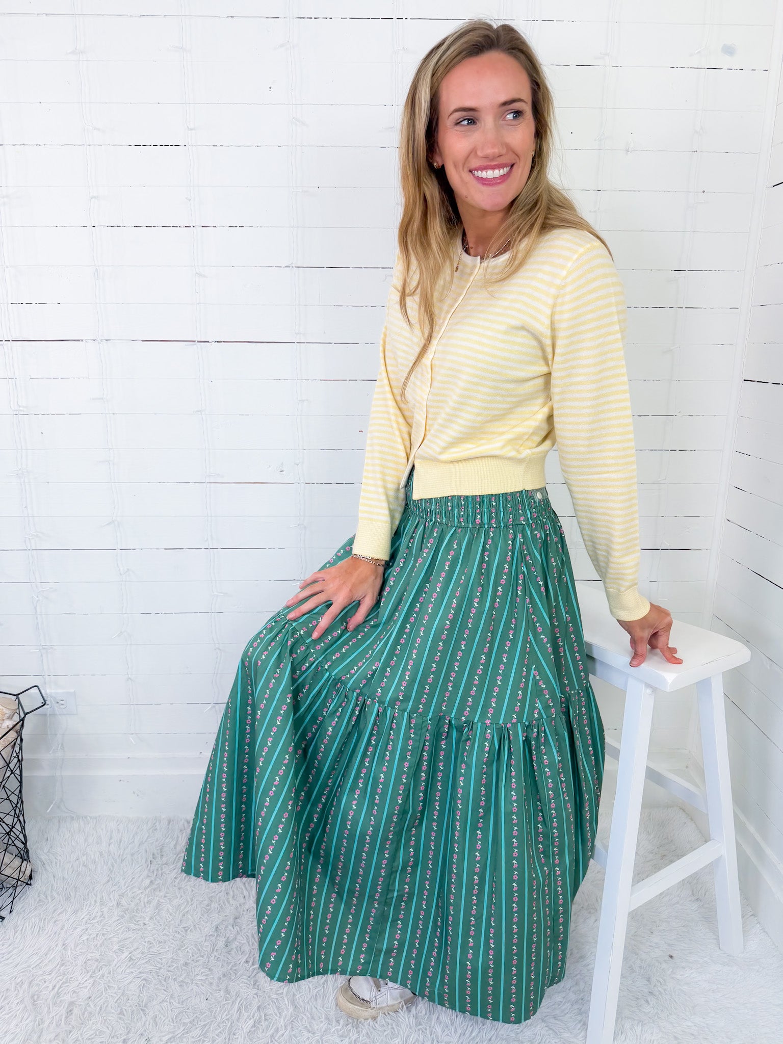 Woman wearing a yellow sweater and green patterned skirt sitting on a white stool against a white wooden paneled wall.