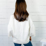 Person wearing a white sweater and blue jeans against a white wooden panel wall.