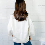Person wearing a white sweater and blue jeans against a white wooden panel wall.