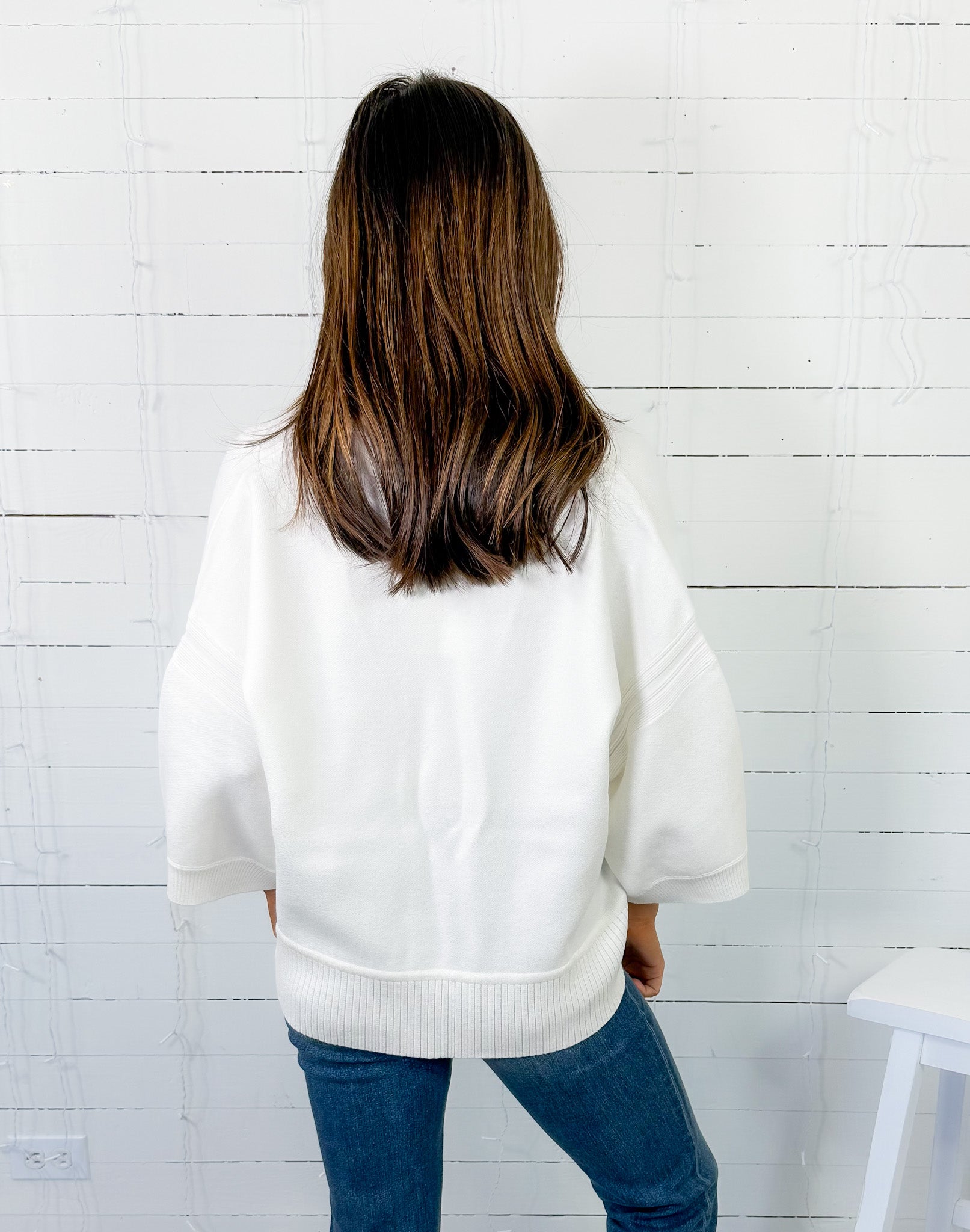 Person wearing a white sweater and blue jeans against a white wooden panel wall.