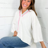 Woman wearing a white hoodie with pink zipper and blue jeans sitting on a white stool against a white wooden wall.