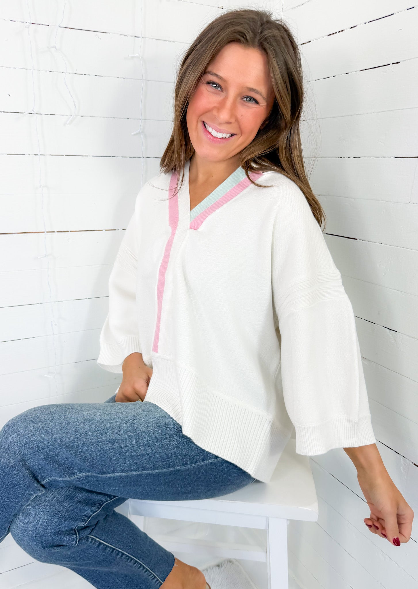 Woman wearing a white hoodie with pink zipper and blue jeans sitting on a white stool against a white wooden wall.