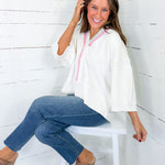 Woman sitting on a white stool wearing a white top with pink accents and blue jeans against a white wooden wall.