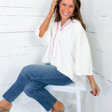 Woman sitting on a white stool wearing a white top with pink accents and blue jeans against a white wooden wall.