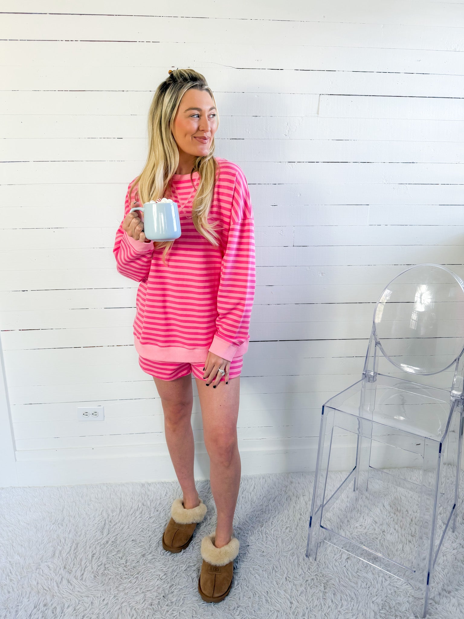Woman in pink striped loungewear holding a mug in a room with a white wall and clear chair.