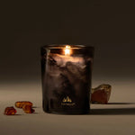 Lit candle in a decorative glass container with amber-colored stones on a dark background