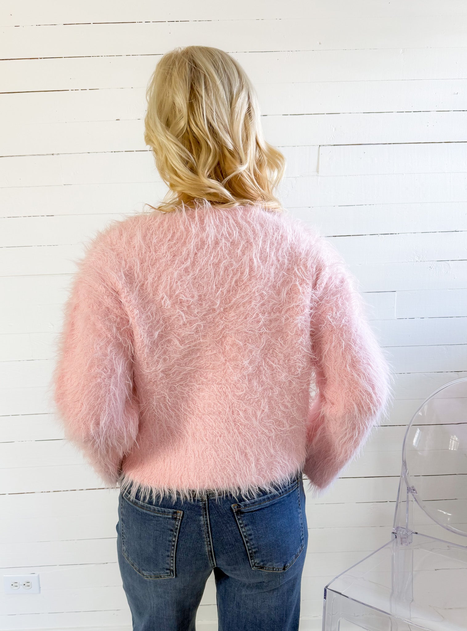 Person wearing a fluffy pink sweater and blue jeans against a white wooden panel background