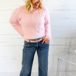 Woman wearing a pink fuzzy sweater and blue jeans standing against a white wooden panel background.