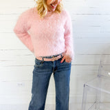 Woman wearing a pink fuzzy sweater and blue jeans standing against a white wooden panel background.