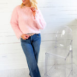 Woman wearing a pink sweater and blue jeans standing next to a clear chair against a white wall.