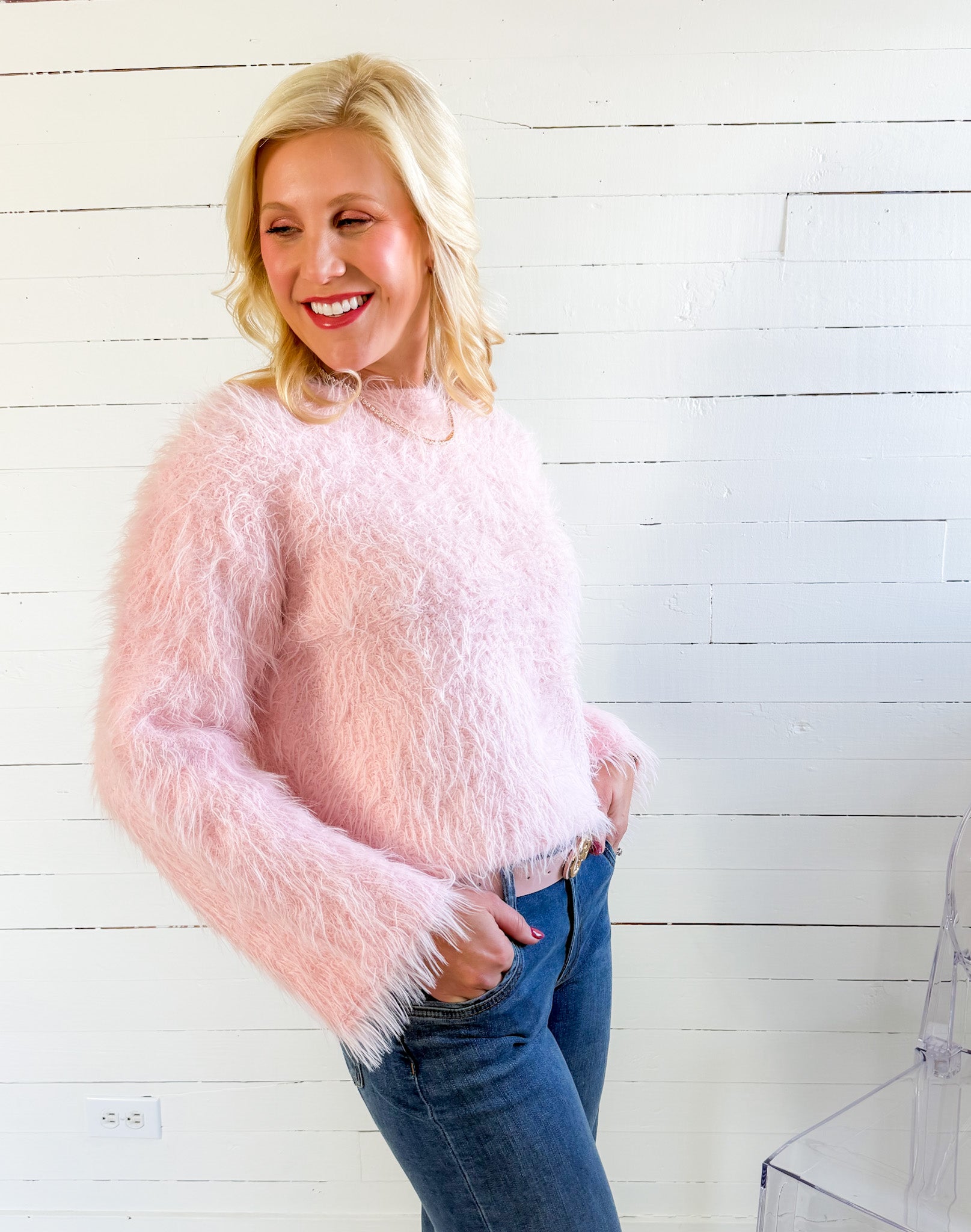 Woman wearing a pink fuzzy sweater and blue jeans against a white wooden panel background