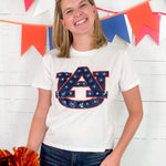 Person wearing a white t-shirt with a logo, holding pom-poms, in front of a colorful banner.