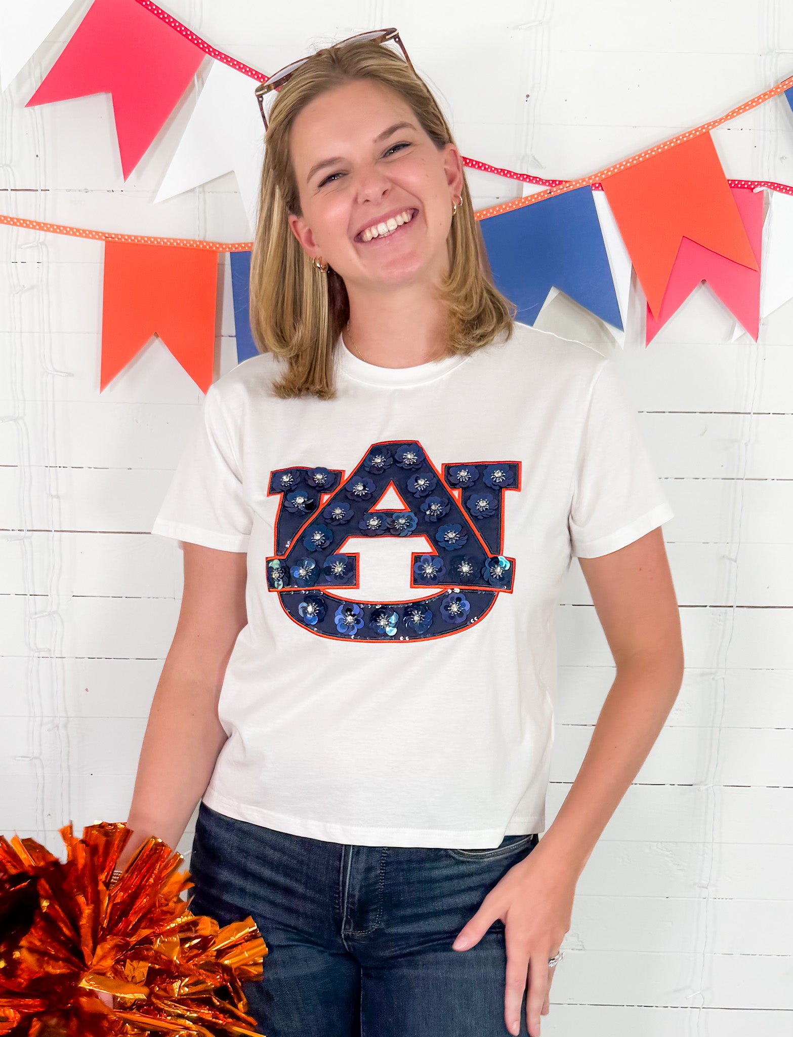 Person wearing a white t-shirt with a logo, holding pom-poms, in front of a colorful banner.