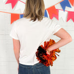 Person holding orange and black pom-poms in front of a colorful flag banner.