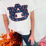 Person wearing a white t-shirt with a large 'AU' logo, holding orange pom-poms against a colorful background.