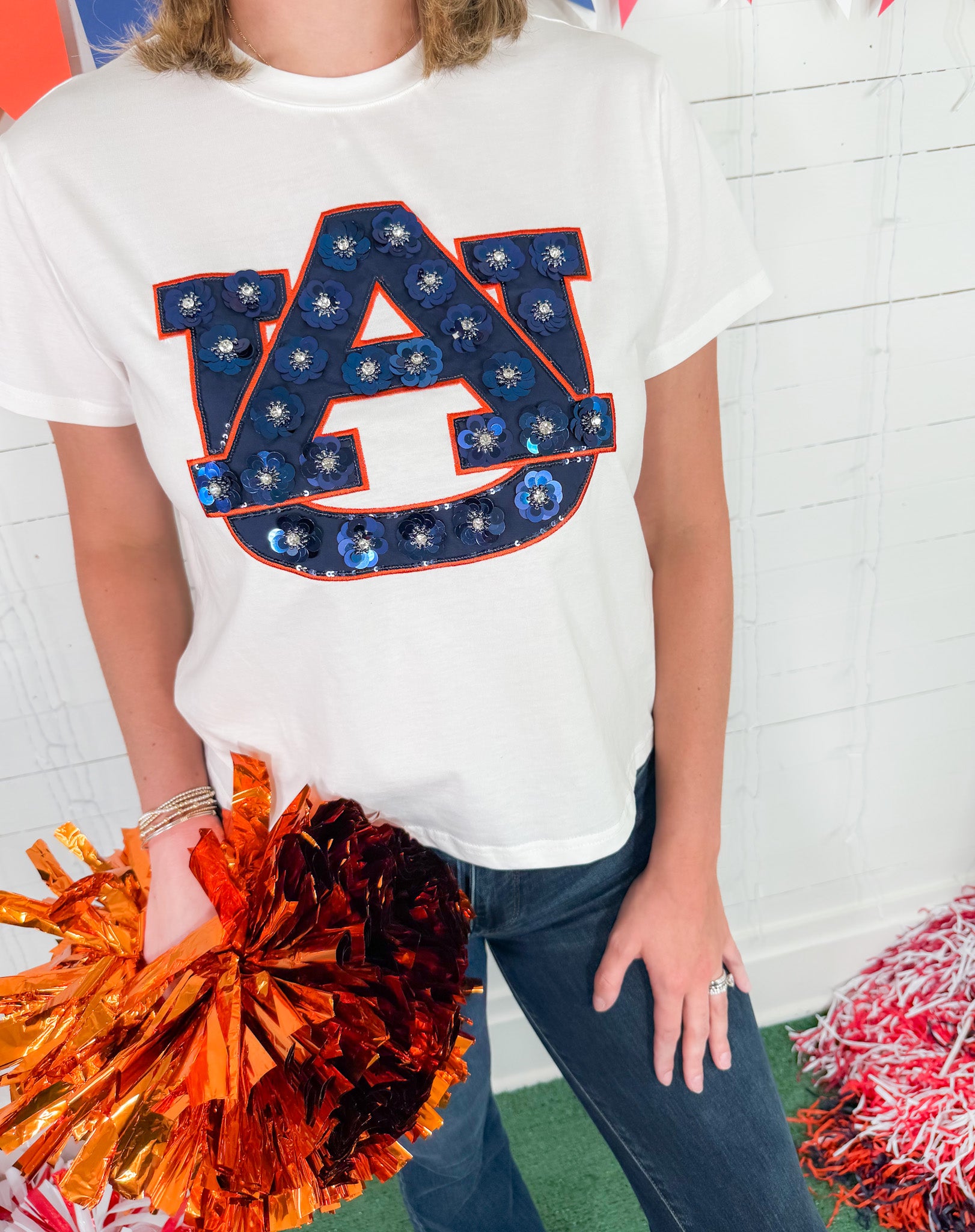 Person wearing a white t-shirt with a large 'AU' logo, holding orange pom-poms against a colorful background.