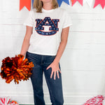 Person wearing a white t-shirt with a logo, holding pom-poms, standing in front of a colorful banner.
