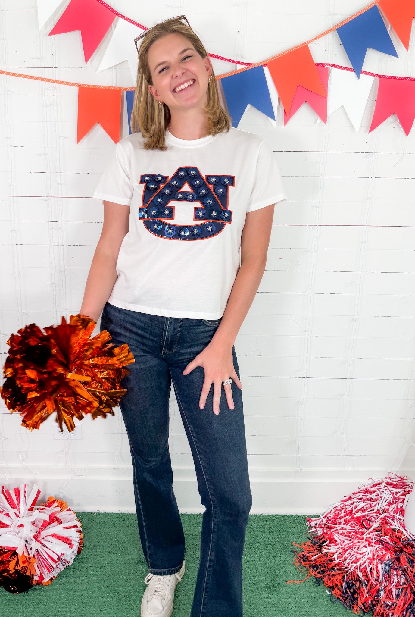 Person wearing a white t-shirt with a logo, holding pom-poms, standing in front of a colorful banner.