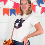 Woman wearing a white t-shirt with a logo, holding pom-poms, in front of a colorful banner.