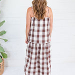 A woman viewed from behind wearing a brown and white gingham check midi dress with spaghetti straps, standing in front of a white shiplap wall next to a potted plant.