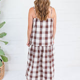 A woman viewed from behind wearing a brown and white gingham check midi dress with spaghetti straps, standing in front of a white shiplap wall next to a potted plant.