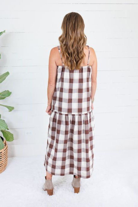 A woman viewed from behind wearing a brown and white gingham check midi dress with spaghetti straps, standing in front of a white shiplap wall next to a potted plant.