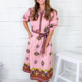 Woman wearing a pink dress with a pattern, standing against a white wooden panel background.