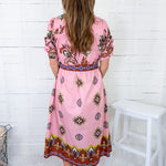 Woman wearing a pink dress with a pattern, standing against a white wooden panel wall.