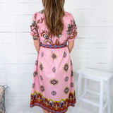Woman wearing a pink dress with a pattern, standing against a white wooden panel wall.